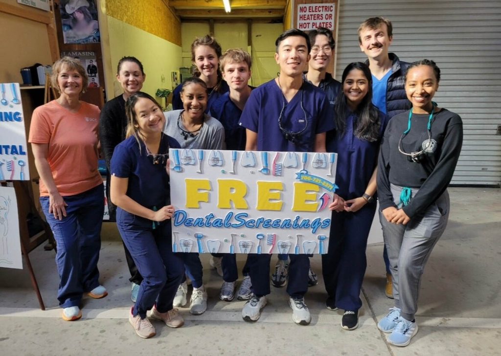 a group of dental students stand in one of the aisle at a flea market, holding a sign that says free dental screenings bordered with plain teeth, teeth with a braces bracket, toothbrushes, and dental cleaning tools, as they smile at the camera.
