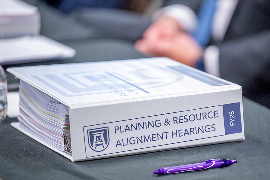 A large binder with the words "Planning and Resource Alignment Hearings" sits on a table.