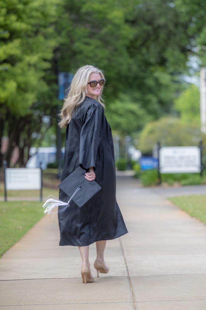 A woman wearing a graduation robe and holding her mortarboard cap walks down a campus sidewalk, away from the camera while looking back over her shoulder