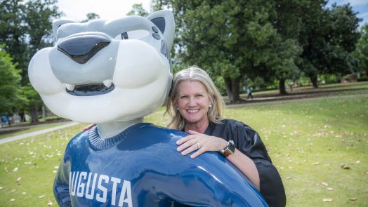 A woman with a cap and gown poses with Augustus statue on a college campus