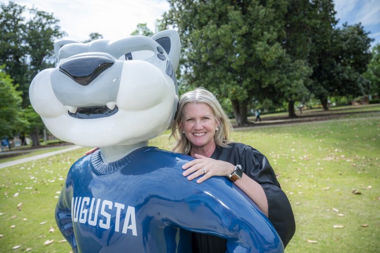 A woman with a cap and gown poses with Augustus statue on a college campus