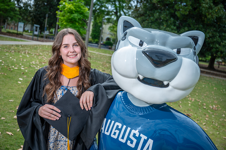 A female college student wears her graduation cap, gown and graduate school hood while posing for a picture with a statue of a college mascot depicting a Jaguar.
