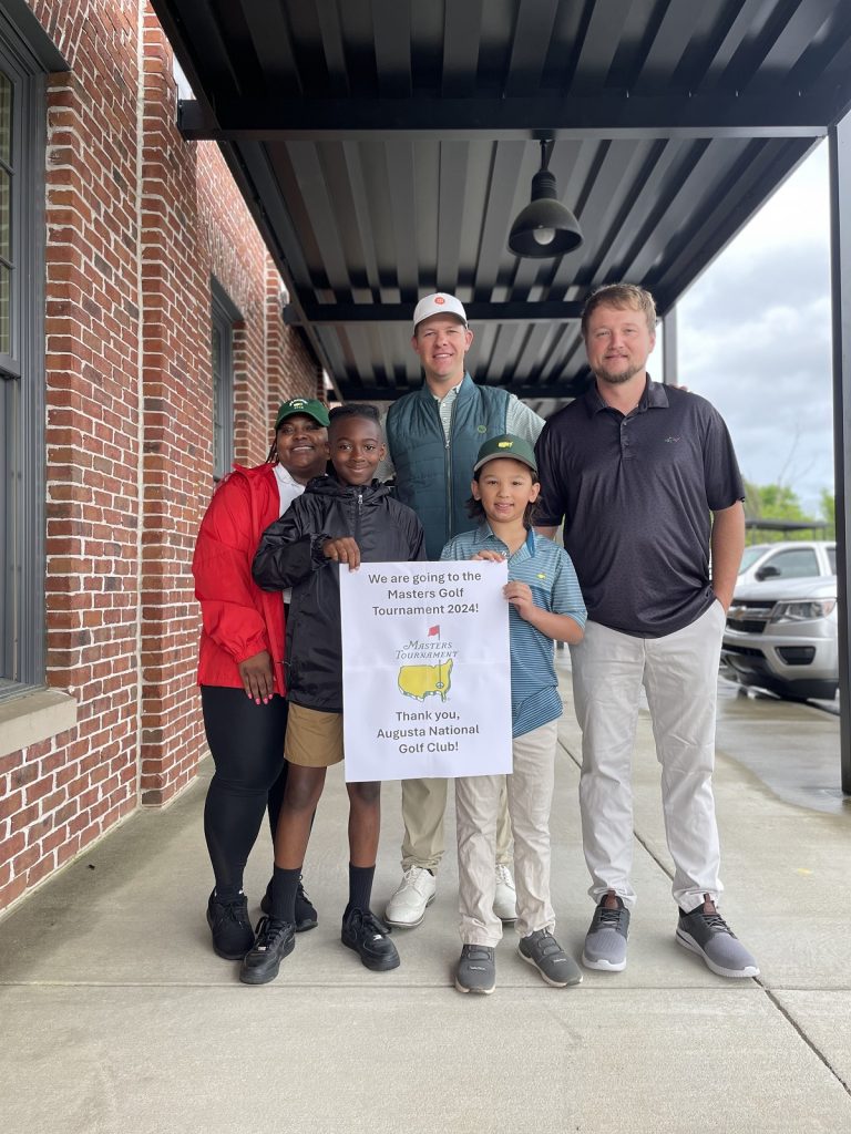 Two men and a woman pose with two young boys outside with a sign that says, "We are going to the Masters Golf Tournament 2024! Thank you, Augusta National Golf Club!"