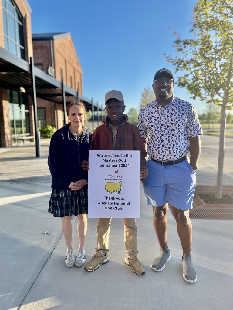 A woman, a man and a young boy pose outside with a sign that says, "We are going to the Masters Golf Tournament 2024! Thank you, Augusta National Golf Club!"