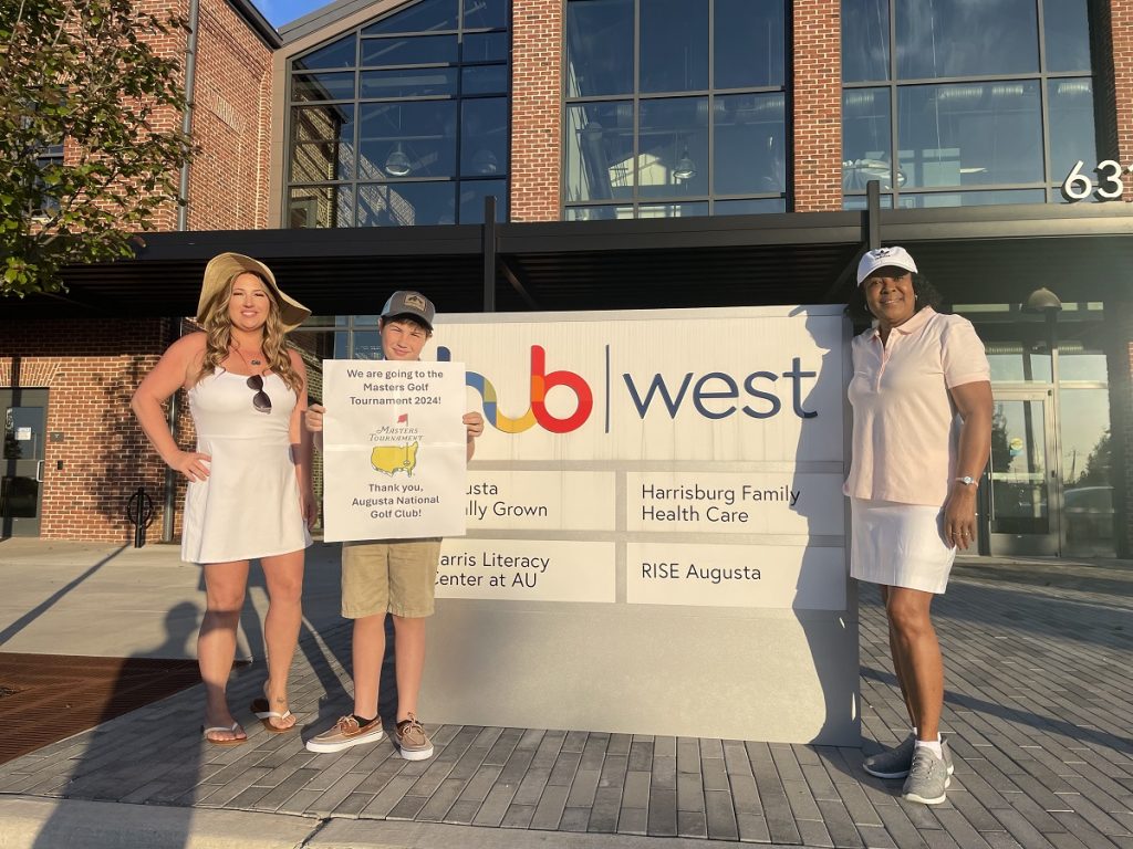 Two women pose with a young boy holding a sign that says, "We are going to the Masters Golf Tournament 2024! Thank you, Augusta National Golf Club!" next to a sign marker for Hub West
