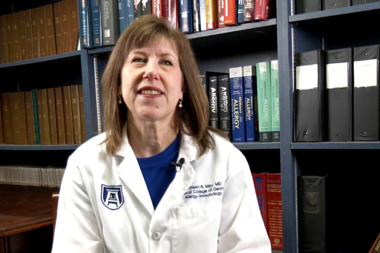 A woman wearing a medical coat sits in front of a large bookcase full of medical texts.