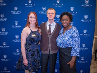 Two women and one man stand in front of a tall backdrop with the logo for Augusta University on it.