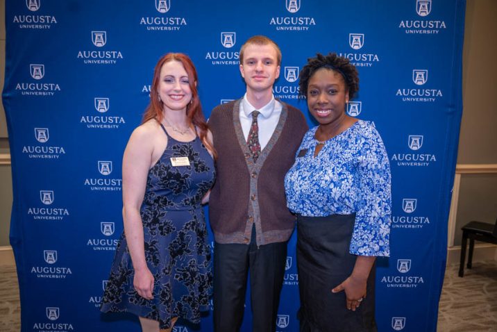 Two women and one man stand in front of a tall backdrop with the logo for Augusta University on it.