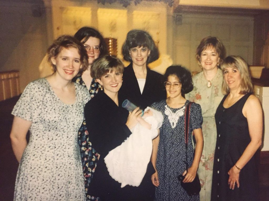 Six women and a teenage girl all in dresses pose in a church a mom bottlefeeding her newborn baby