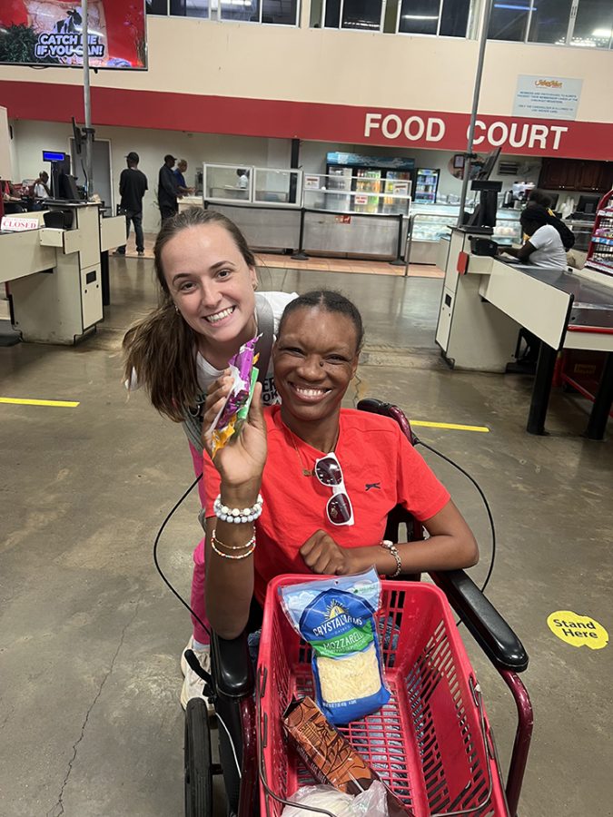 a woman in a red shirt with white sunglasses hanging from her shirt sits in a wheelchair smiling with a basket containing groceries on her lap while showing off one item and another woman pushing her crouches down to her level to smile for a photo in a local market
