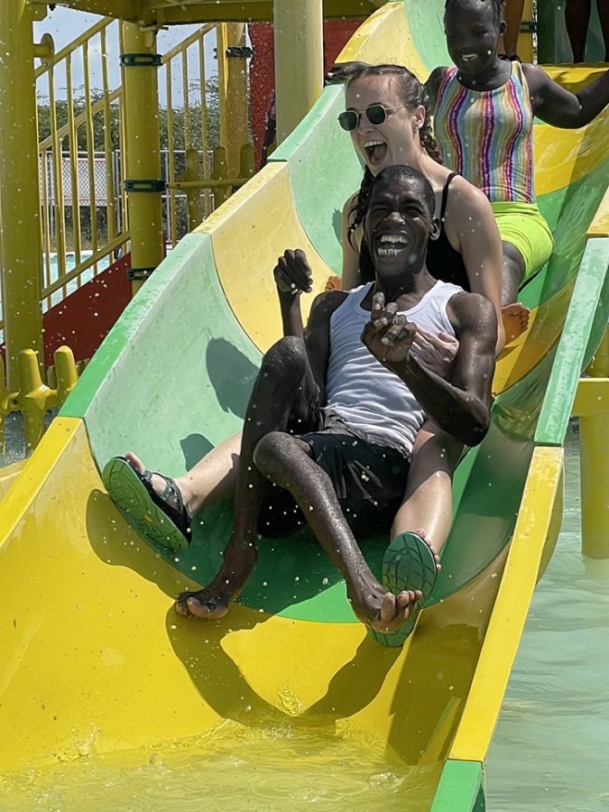 A female student helps a male resident slide down a green and yellow waterslide while another resident slides down behind them