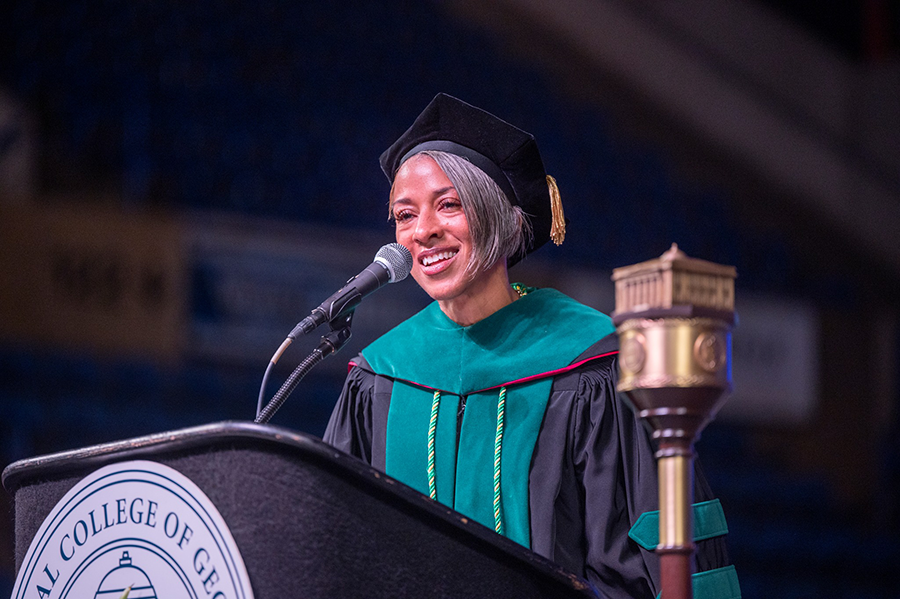 A woman wearing full college graduation regalia of cap, gown and hood stands at a podium with a microphone to address a large audience.