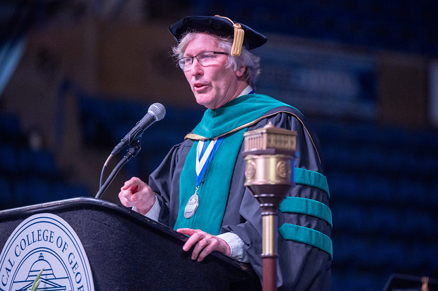 A man wearing full college graduation regalia of cap, gown and hood stands at a podium with a microphone to address a large audience.