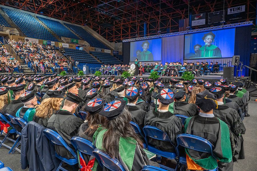 Recent medical school graduates sit in a large arena listening as a ceremony is held.