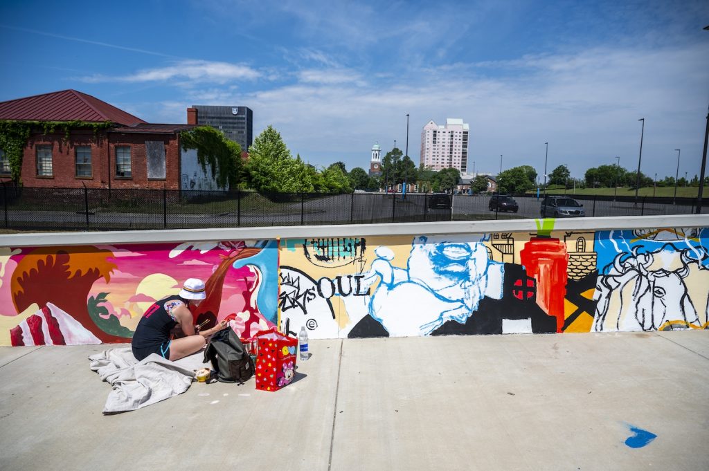 A woman paints a mural on a wall of a pedestrian bridge next to another mural.