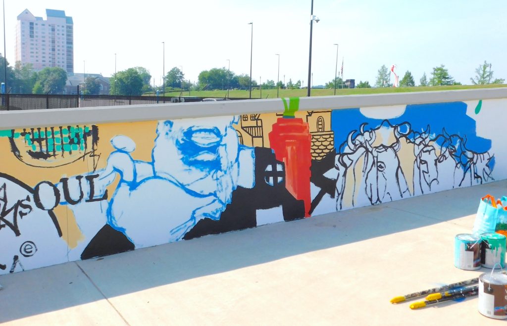 A partially finished mural is painted on a wall of a pedestrian bridge.