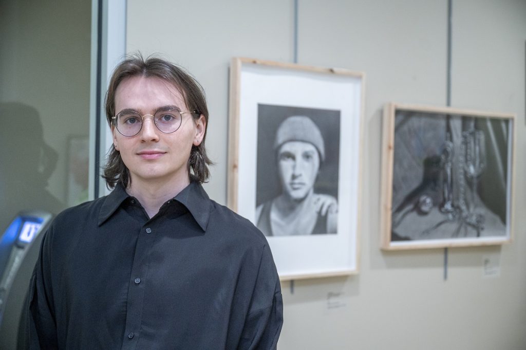 A male college student poses for a picture in front of several pieces of art he created.