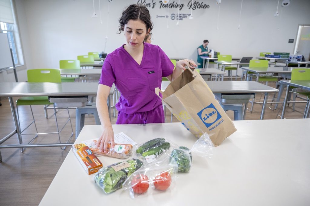 A female medical student in scrubs places fresh vegetables and other foods into a paper bag.
