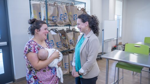 A woman holds a baby while speaking with another woman in a large commercial kitchen.