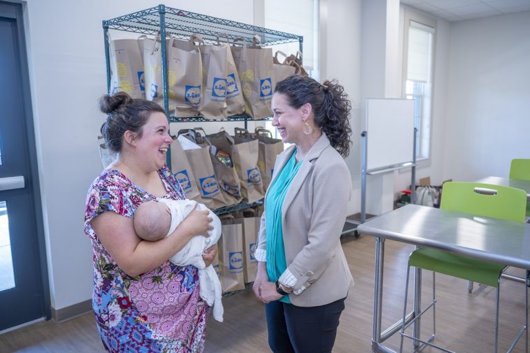 A woman holds a baby while speaking with another woman in a large commercial kitchen.