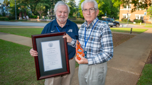 Two men stand outside. One holds a framed award while the other holds a colorful glass trophy in the shape of a vase.