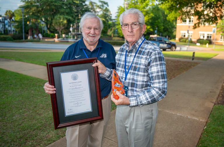 Two men stand outside. One holds a framed award while the other holds a colorful glass trophy in the shape of a vase.