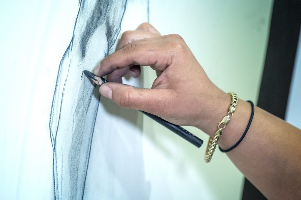 A woman's hand holds a charcoal pencil to a sketchpad while drawing a human leg in great detail.