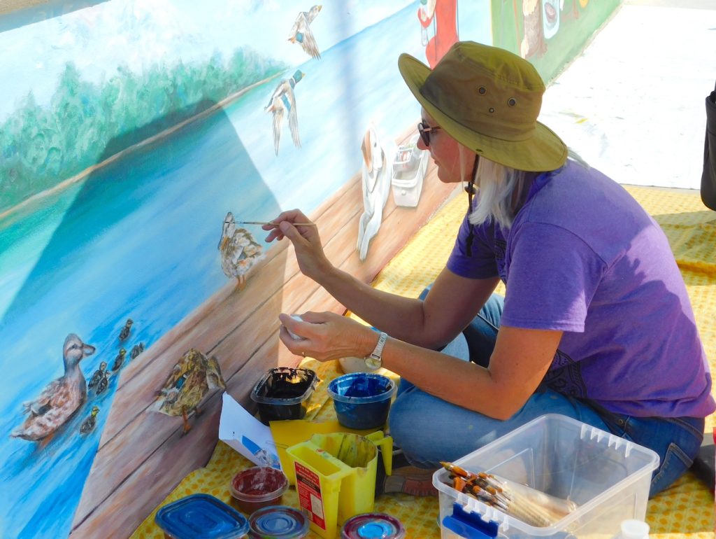 A woman paints a duck as part of a mural depicting a peaceful dock scene.