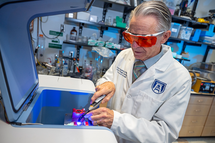 Man wearing a scientific lab coat and protective glasses uses a piece of lab equipment in the form of a pen with a high-powered light on the end.