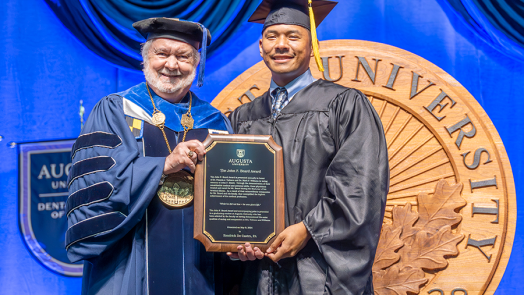 A man wearing traditional graduation cap and gown hands a college graduate, also wearing a graduation cap and gown, a plaque for an award during a graduation ceremony.