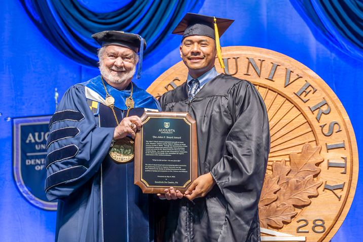 A man wearing traditional graduation cap and gown hands a college graduate, also wearing a graduation cap and gown, a plaque for an award during a graduation ceremony.