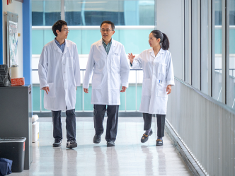 Two men and a woman, all wearing lab coats, walk down a hallway in a research building.