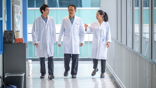 Two men and a woman, all wearing lab coats, walk down a hallway in a research building.