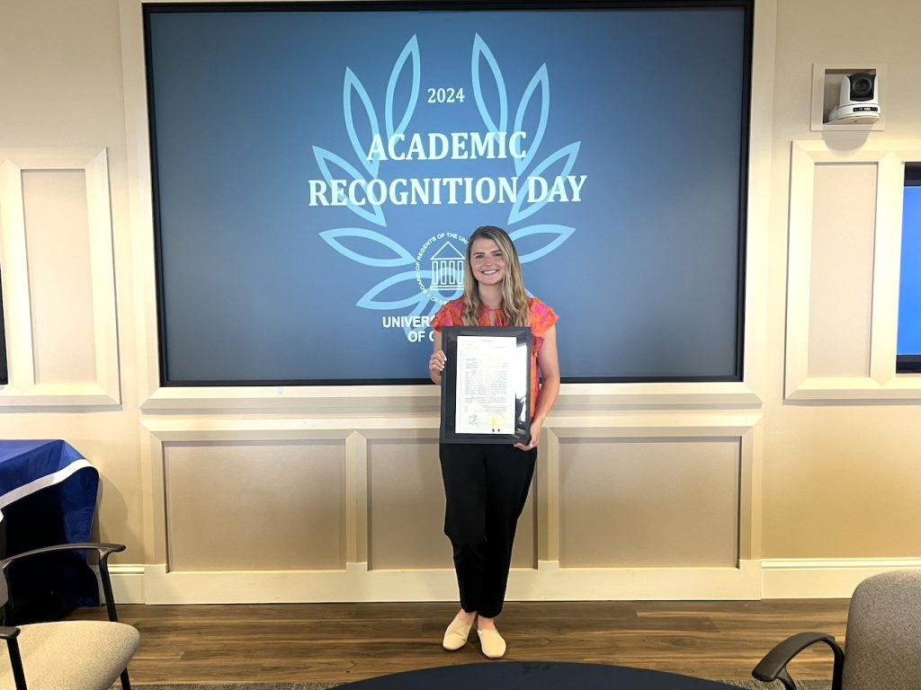 A woman stands in front of a projection screen holding an award. Behind her, the words "2024 Academic Recognition Day" with the logo for the University System of Georgia are displayed.