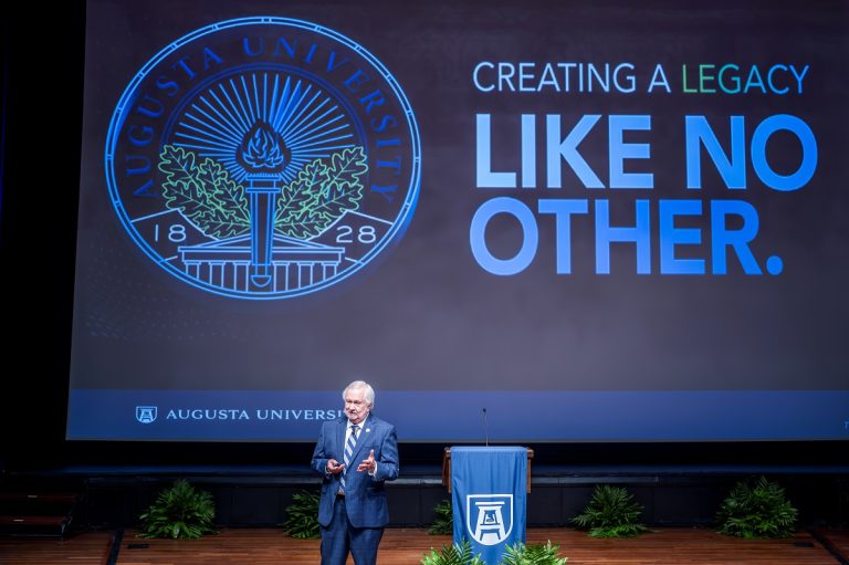 A man stands on a stage in a large auditorium and addresses a large crowd.