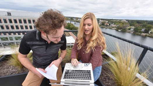 Two college students look at the screen of a laptop while sitting outside.