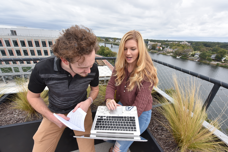 Two college students look at the screen of a laptop while sitting outside.