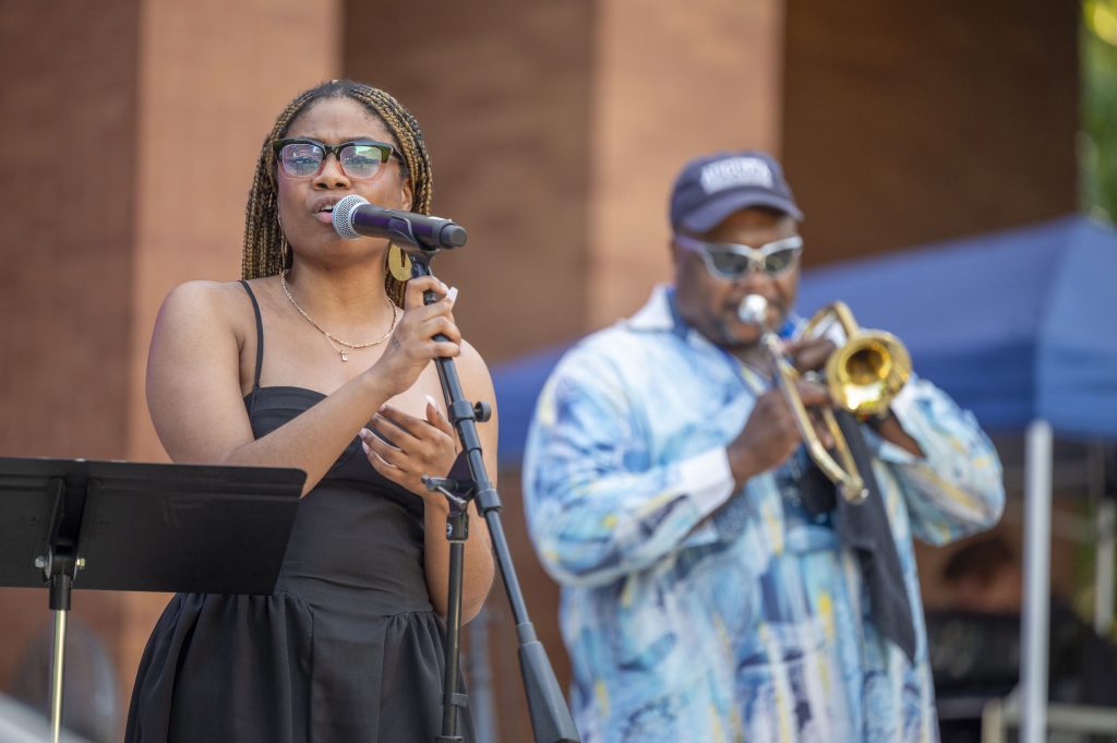 A woman sings into a microphone while a man plays trombone in background.