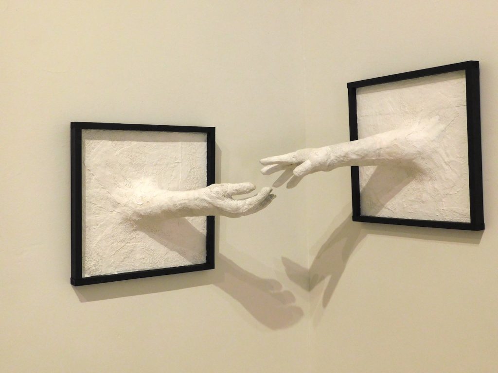 Artwork made of plaster hung on a wall depicts two hands reaching out towards each other.