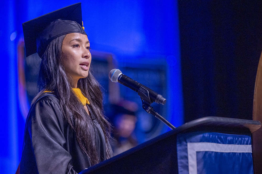 A female college graduate wearing ca and gown stands at a podium with a microphone and sings to a crowd.