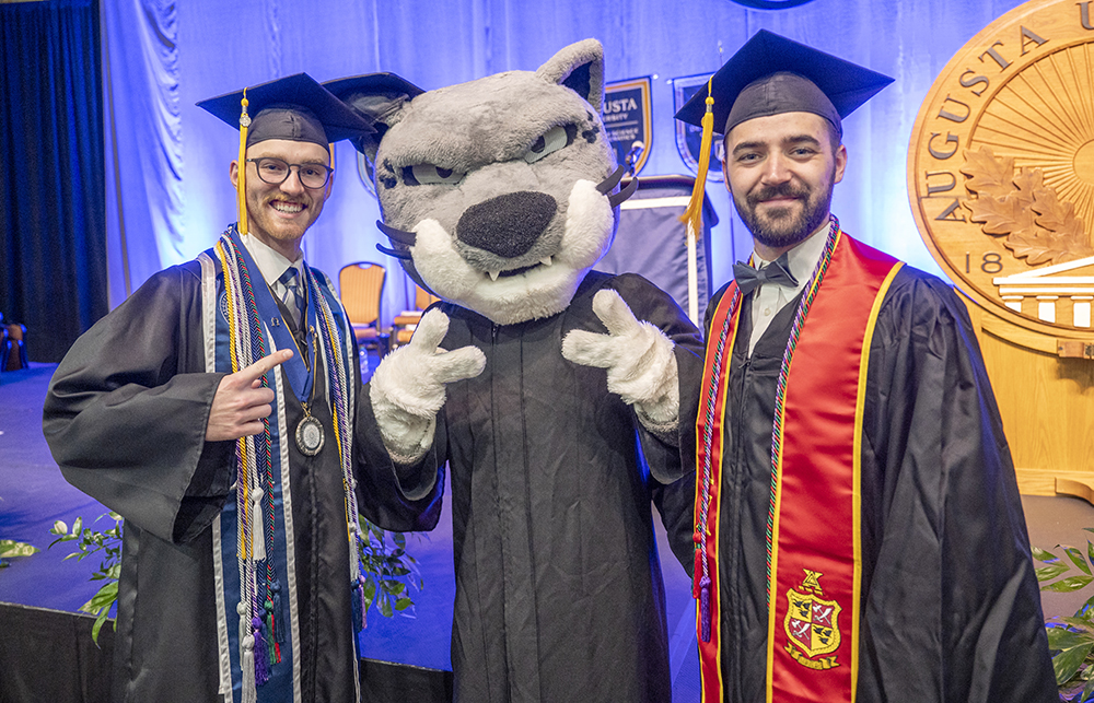 Two male college graduates wearing cap and gown stand with a jaguar college mascot, also in cap and gown.