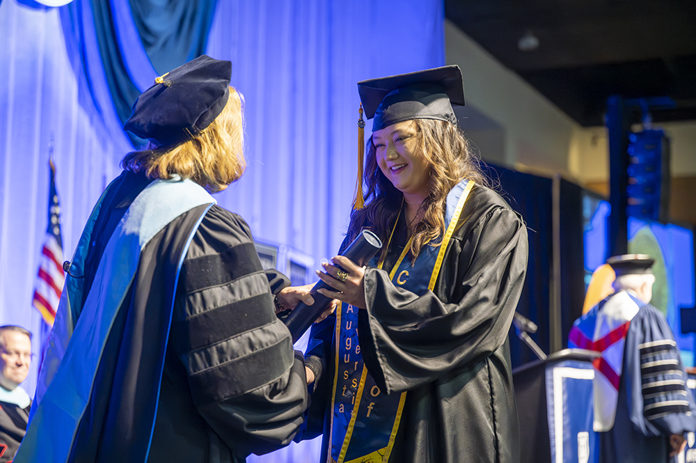 A woman in graduation cap and gown shakes hands with a female college graduate while handing her a diploma.