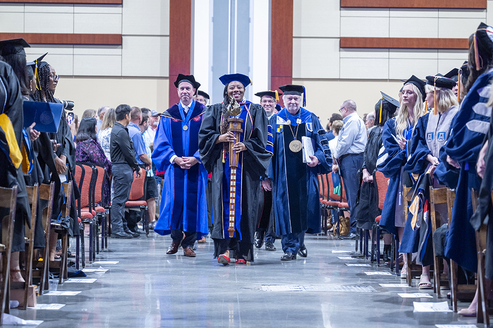 Faculty of a university, all wearing full graduation regalia of cap and gown, walk down a center aisle during a graduation ceremony.
