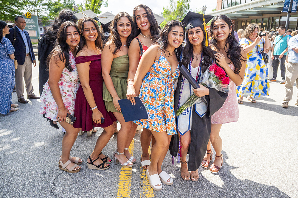 Six college women huddle around a female college graduate in full graduation cap and gown to celebrate her graduation.