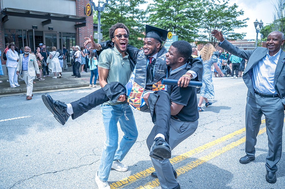 Two college men hold a friend up in celebration of him graduating from college.