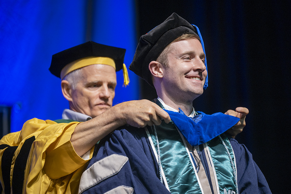 A man wearing college graduation cap, gown and hood places a new hood over the head of a recent college graduate, also wearing cap and gown.