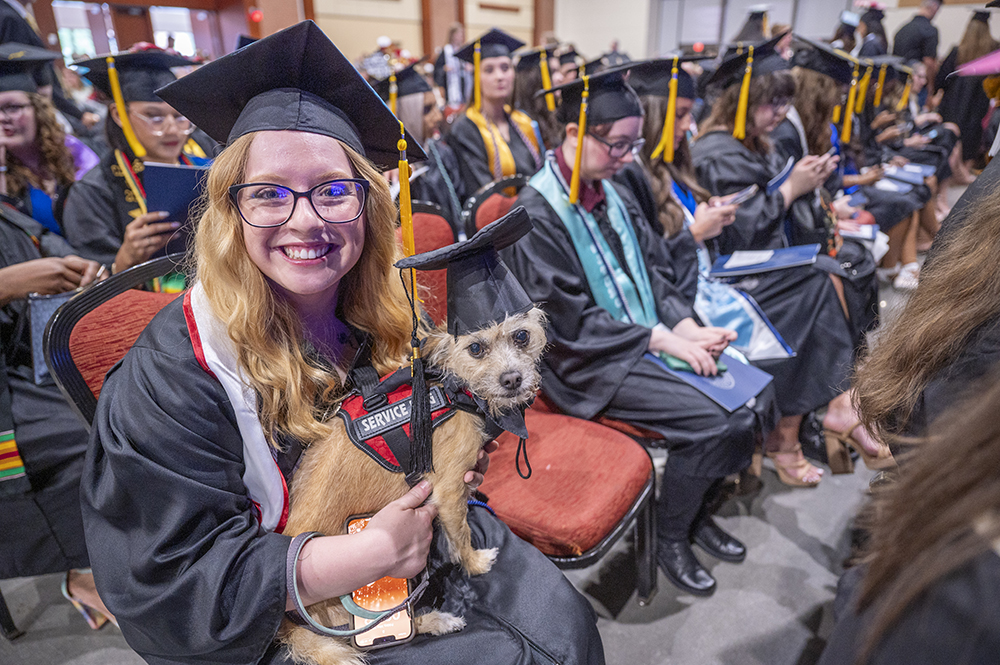 A female college graduate wearing cap and gown sits while holding her service dog.
