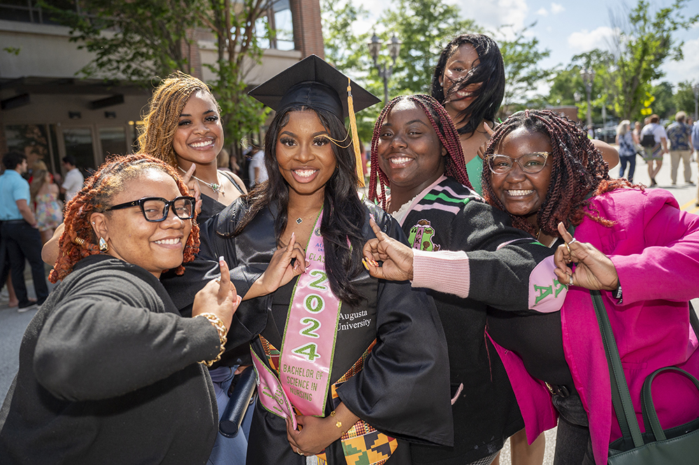 Five women gather around a female college graduate wearing cap and gown to celebrate her graduation.