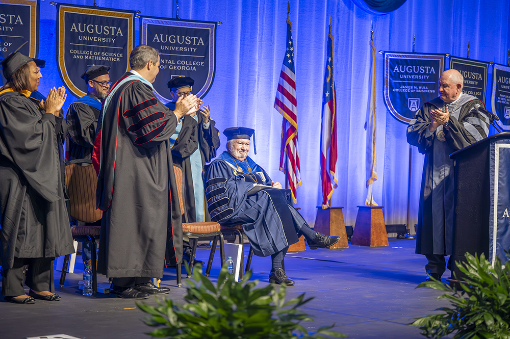 An older gentleman wearing graduation cap and gown sits on a stage while others around him offer a standing ovation.