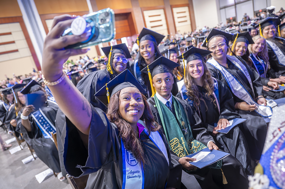 A group of college graduates, all wearing graduation cap and gown, pose for a selfie as one woman holds her phone up.